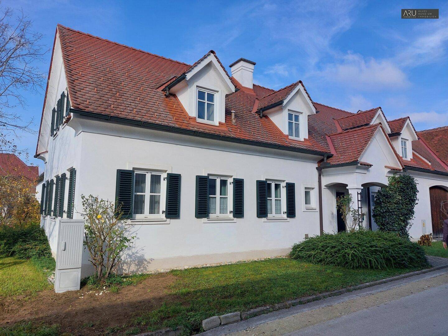 Seitenansicht eines traditionellen Landhauses mit weißer Fassade, grünen Fensterläden und rotem Ziegeldach.