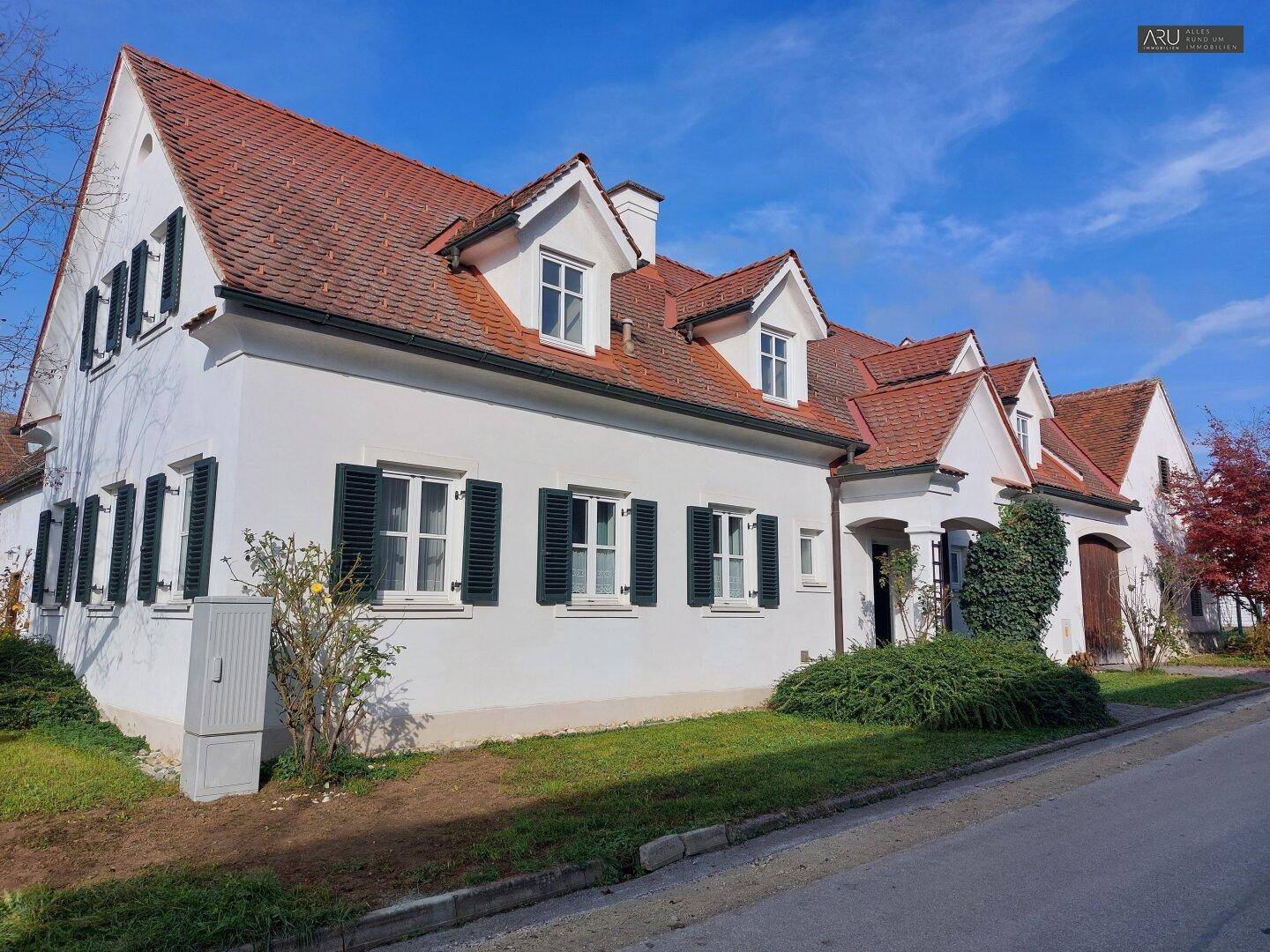 Seitenansicht des Landhauses mit weißer Fassade, grünen Fensterläden und rotem Ziegeldach unter blauem Himmel.