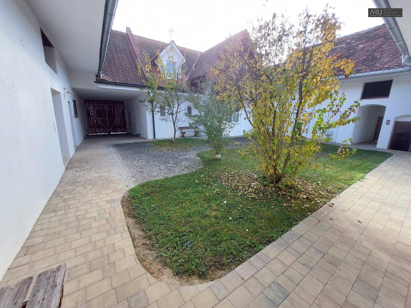 Sonniger Innenhof mit gepflastertem Bereich, Rasenflächen und jungen Bäumen, umgeben von Gebäuden.