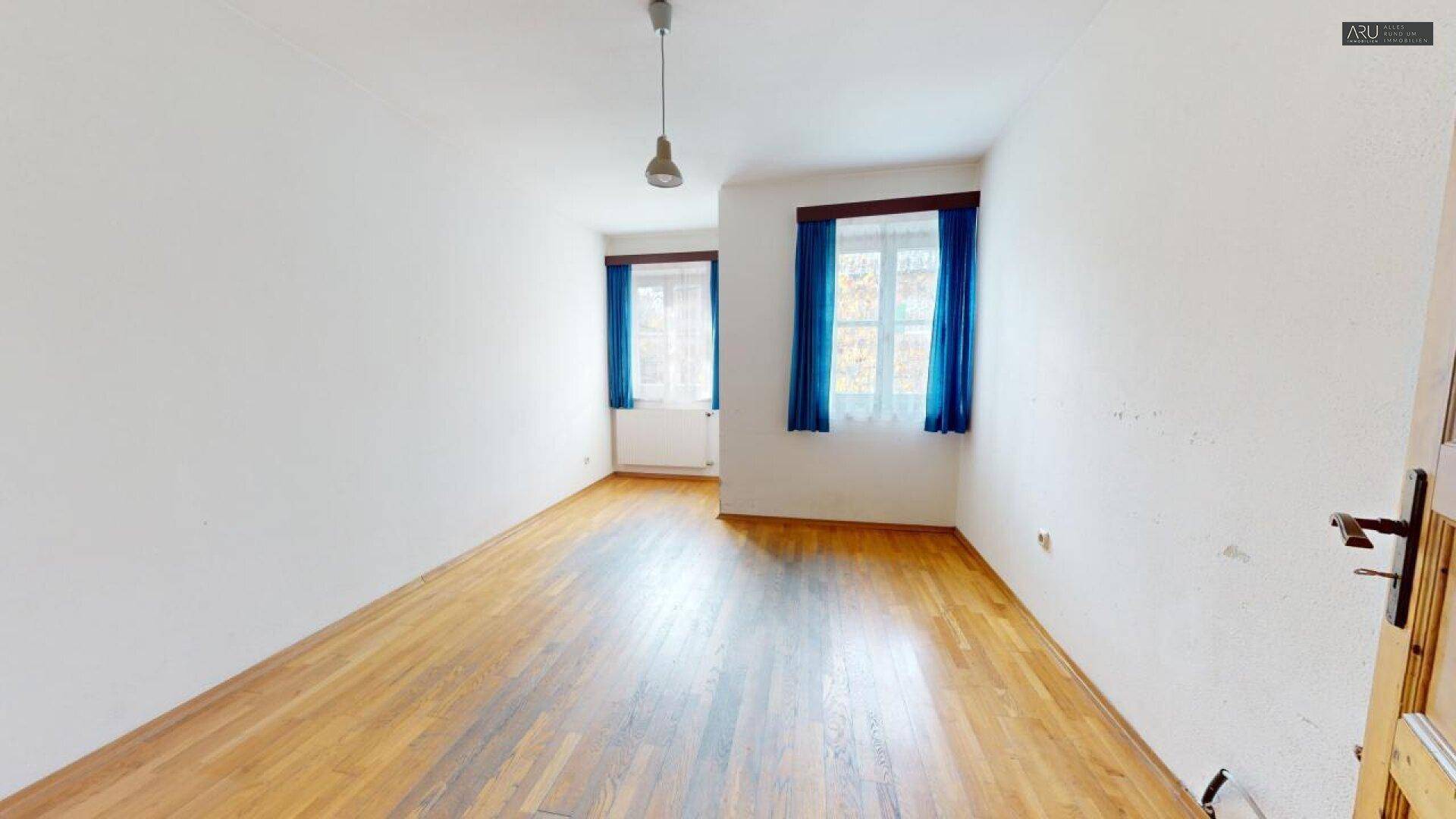 Geräumiges Zimmer mit Holzboden, weißen Wänden und zwei Fenstern mit blauen Vorhängen.