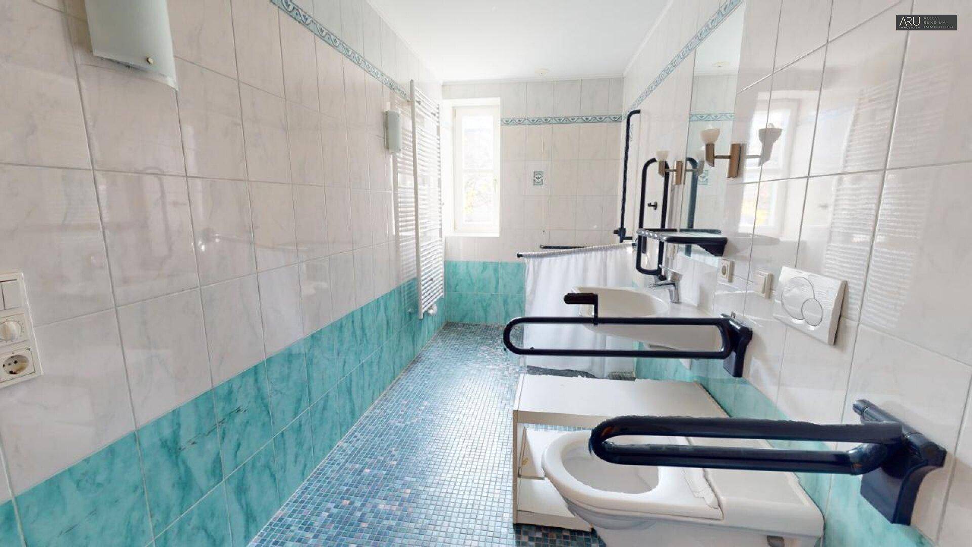 Barrierefreies Badezimmer mit hellblauen Fliesen, Duschbereich und Haltegriffen an der Toilette.