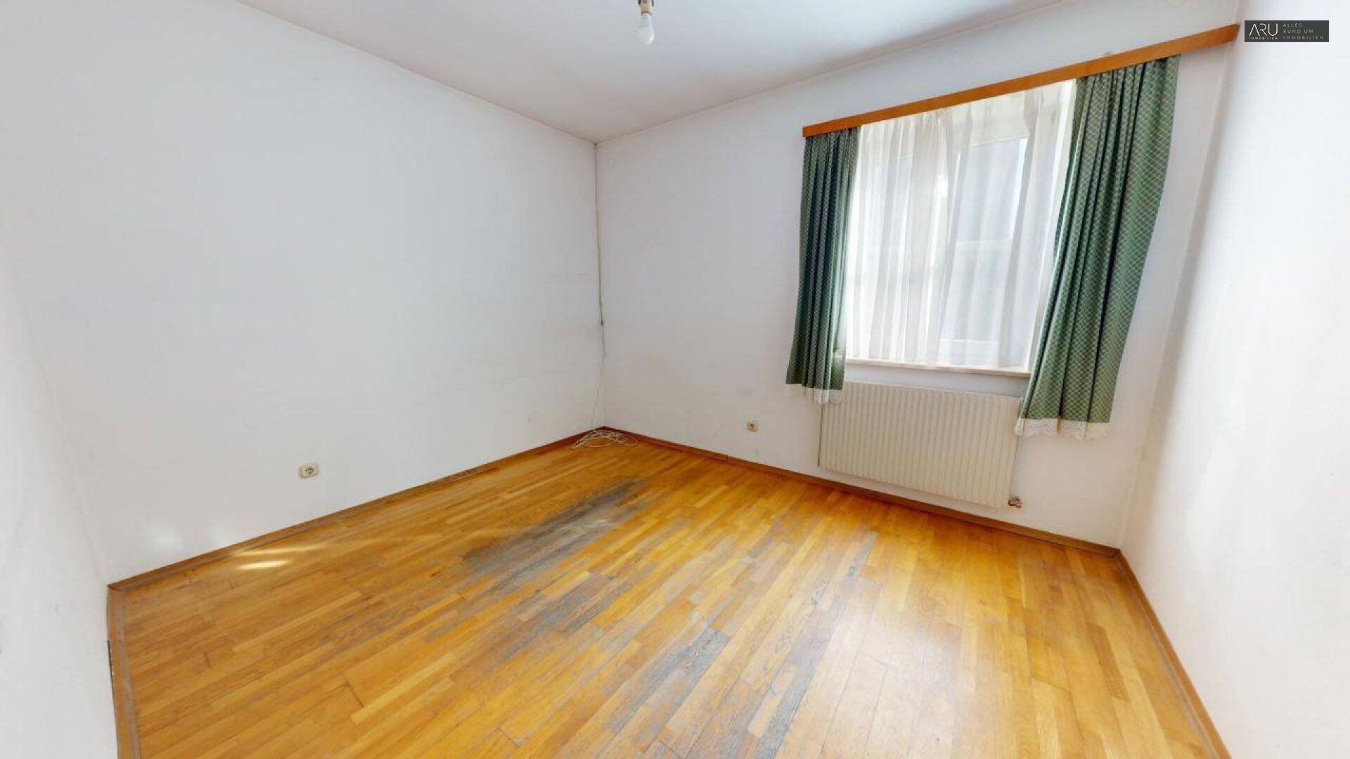Helles Zimmer mit Holzboden, weißen Wänden und einem Fenster mit grünen Vorhängen.