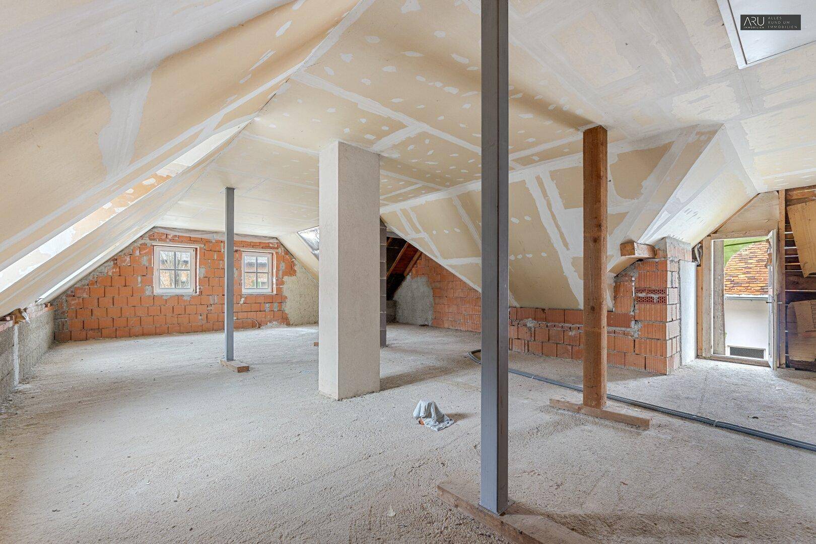 Geräumiger unausgebauter Dachboden mit Ziegelwänden, Stützpfeilern und Fenstern in den Dachschrägen.