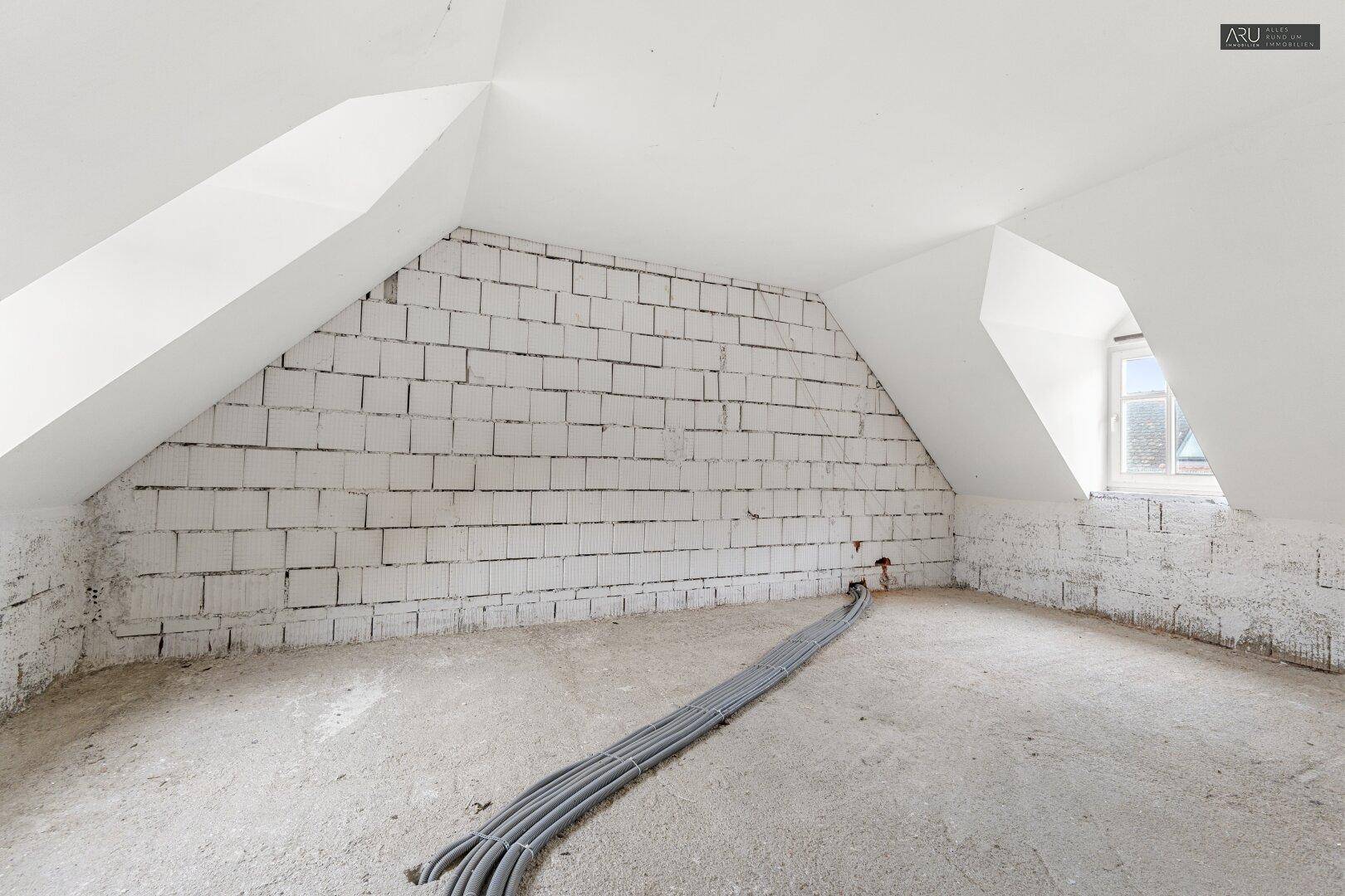 Unausgebauter Dachbodenraum mit weiß gestrichenen Ziegelwänden, schrägen Decken und einem kleinen Fenster.