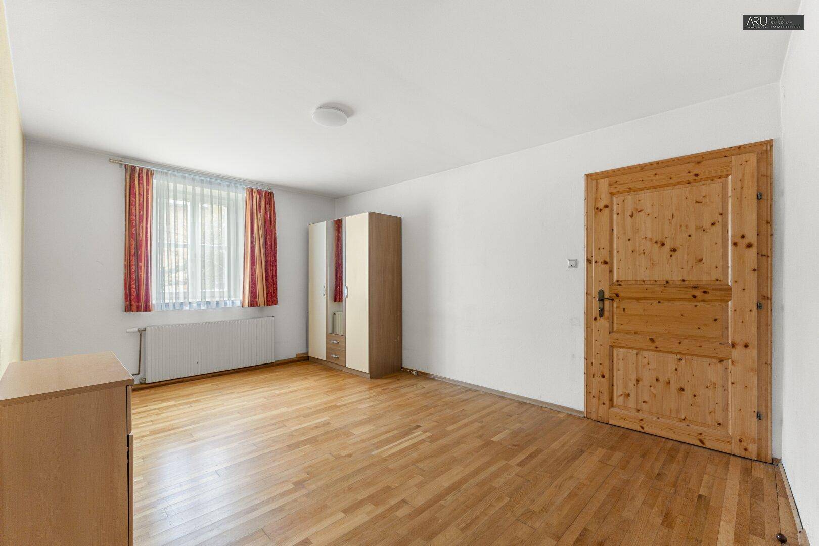 Helles Schlafzimmer mit Holzboden, weiß gestrichenen Wänden und einem Fenster mit Vorhängen.