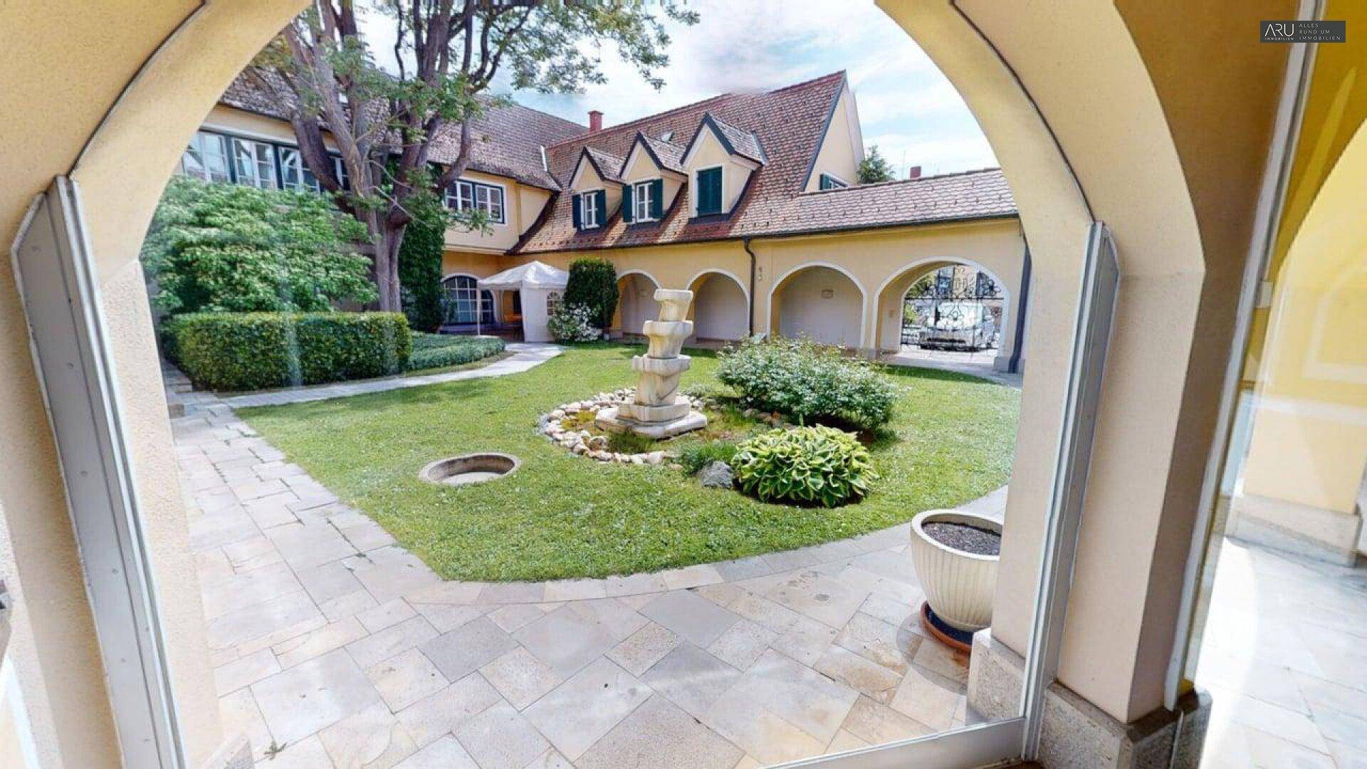 Ein gepflegter Innenhof mit einem dekorativen Brunnen, umgeben von Rasenflächen und einem Gebäude mit Arkaden.