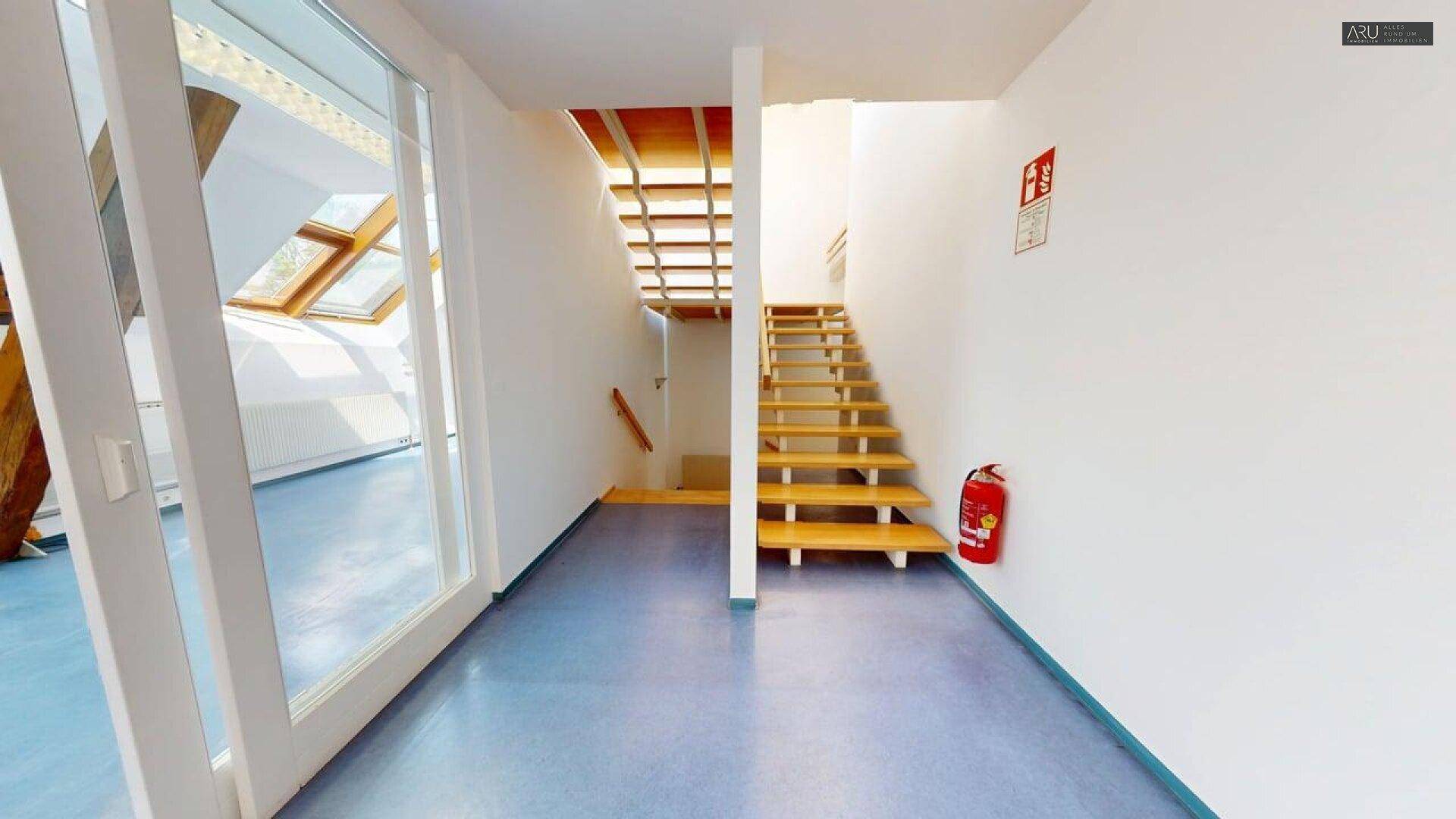 Heller Flur mit blauer Bodenfläche, Holztreppe und großen Fenstern, die Tageslicht hereinlassen.