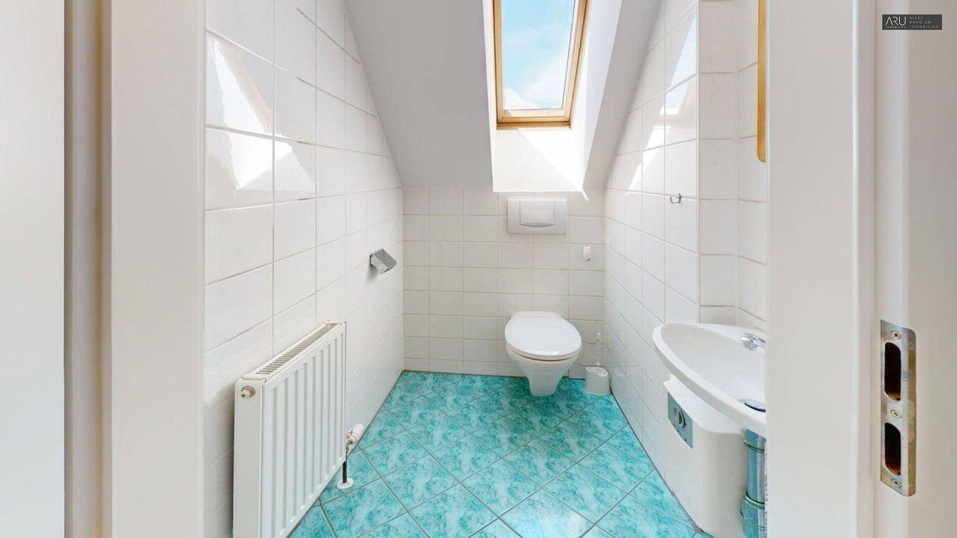 Kleine Toilette im Dachgeschoss mit weiß gefliesten Wänden, Dachfenster und blauem Boden.