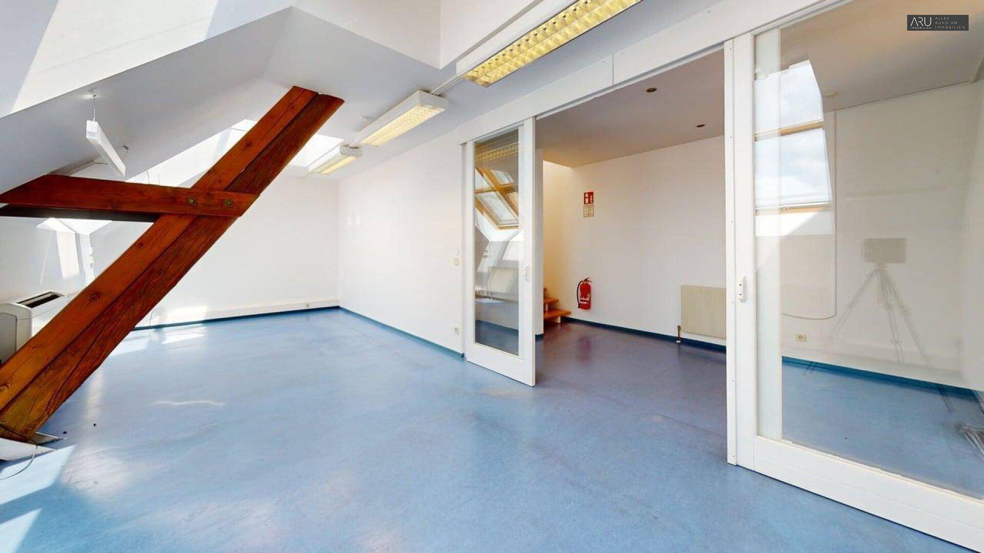 Büroraum mit blauer Bodenfläche, sichtbaren Holzbalken und Glasschiebetüren.
