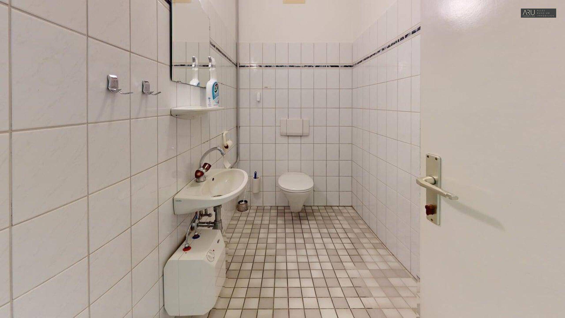 Geräumige Toilette mit weißen Wandfliesen, kleinem Waschbecken und quadratischen Bodenfliesen.