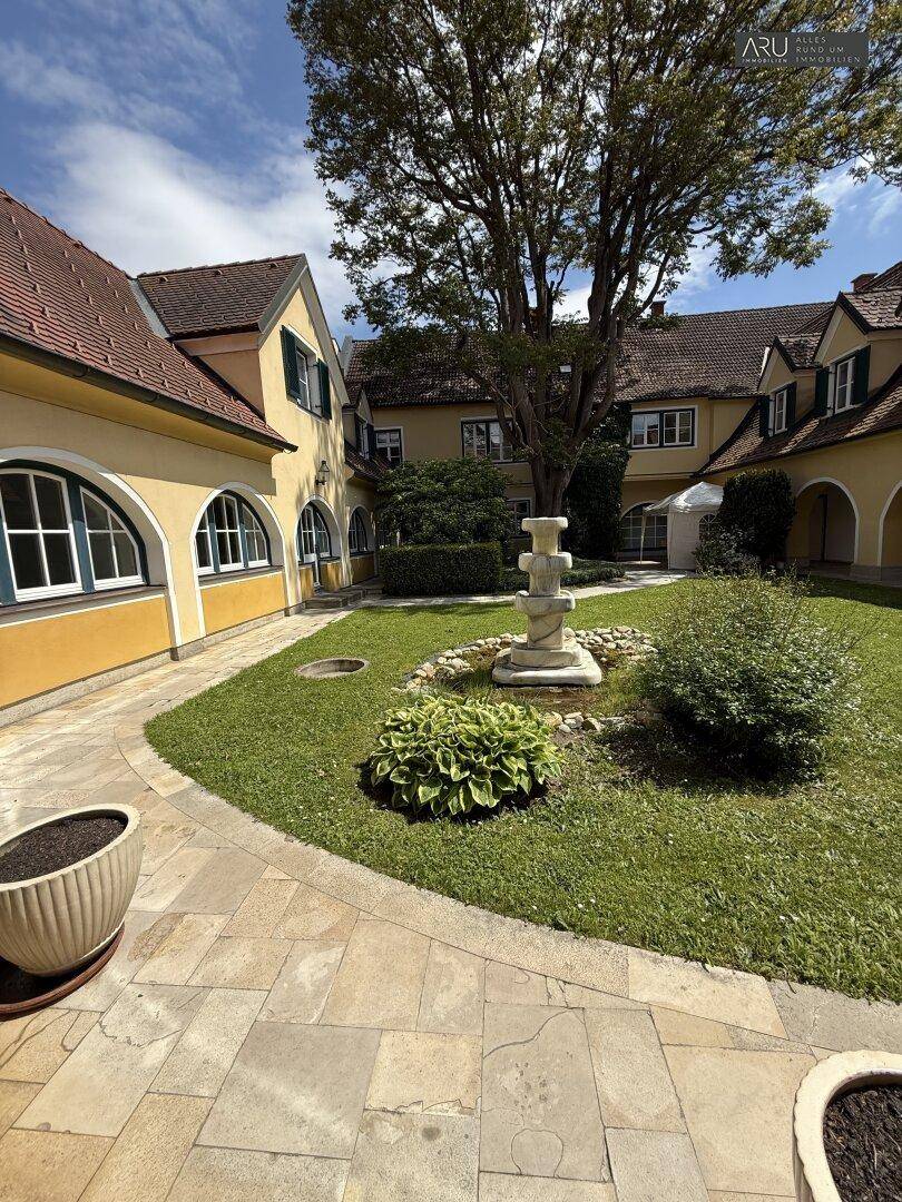 Großer Innenhof mit altem Baum, Brunnen und gepflasterten Wegen.