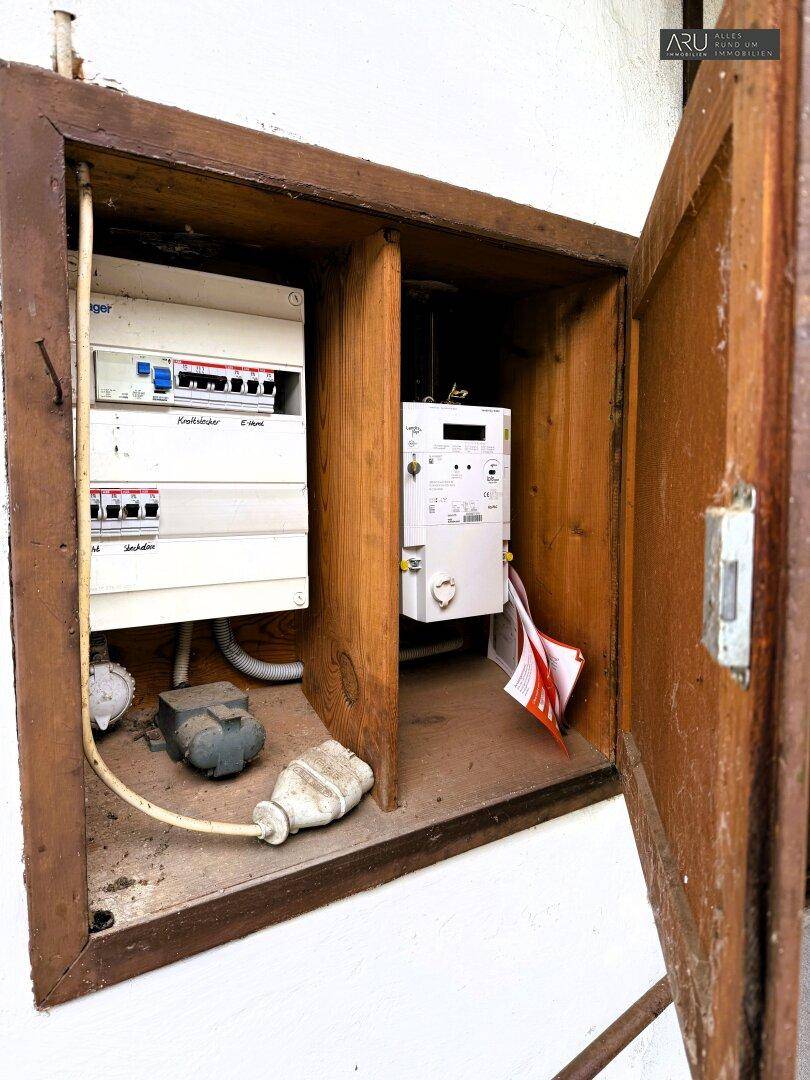 Offener Sicherungskasten und Stromzähler in einem Holzschrank, der die elektrische Installation zeigt.