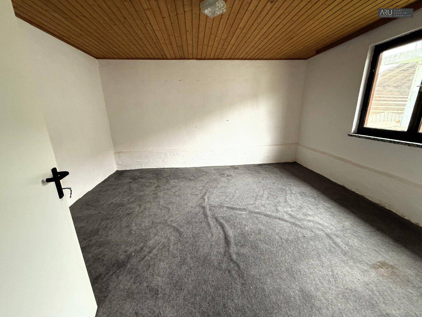 Leeres Zimmer mit grauem Teppichboden, weißen Wänden und einer Holzdecke, bereit zur Einrichtung.