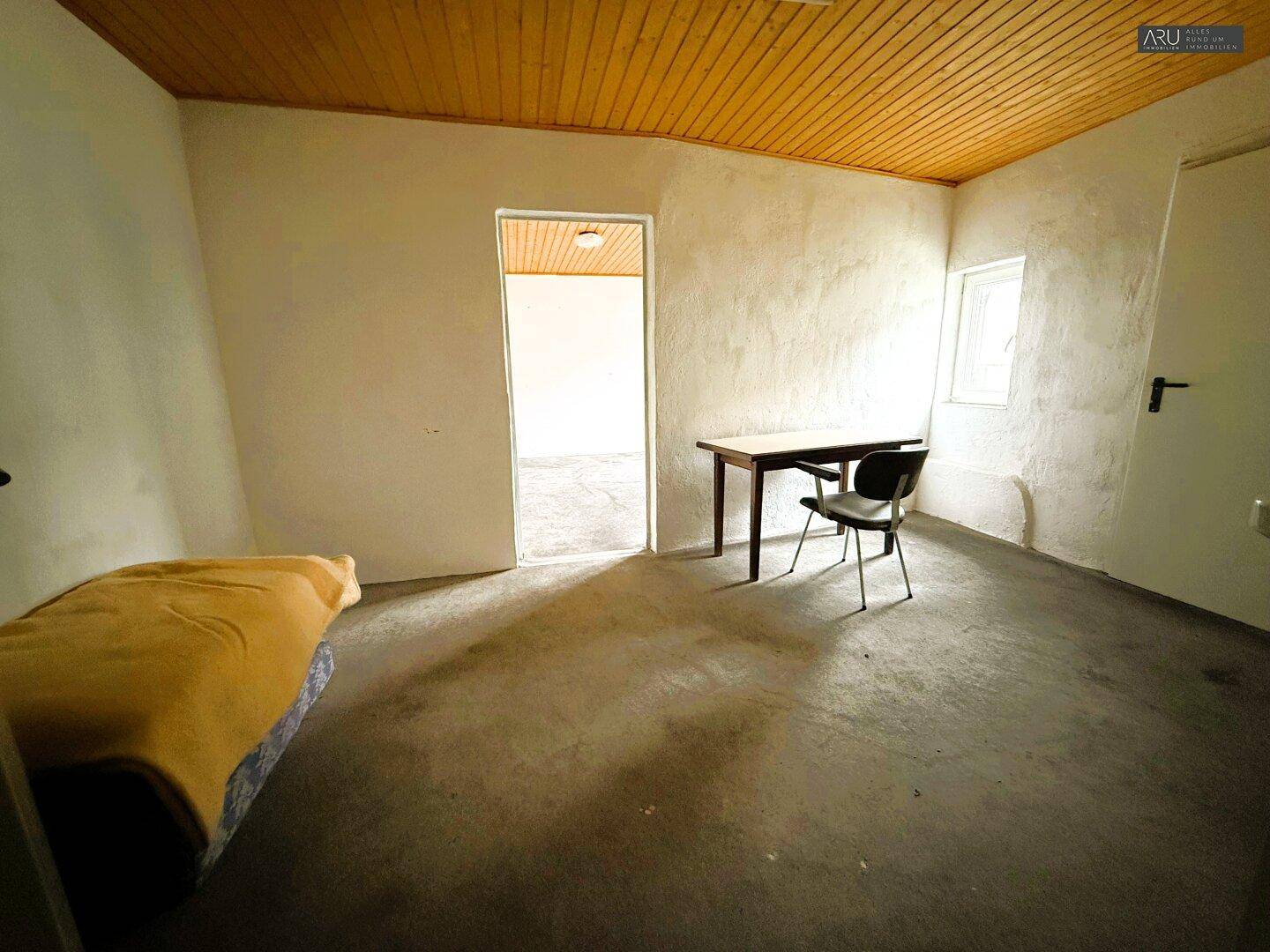 Helles Zimmer mit Betonboden, weißen Wänden und einer Holzdecke, ausgestattet mit einem einfachen Tisch.