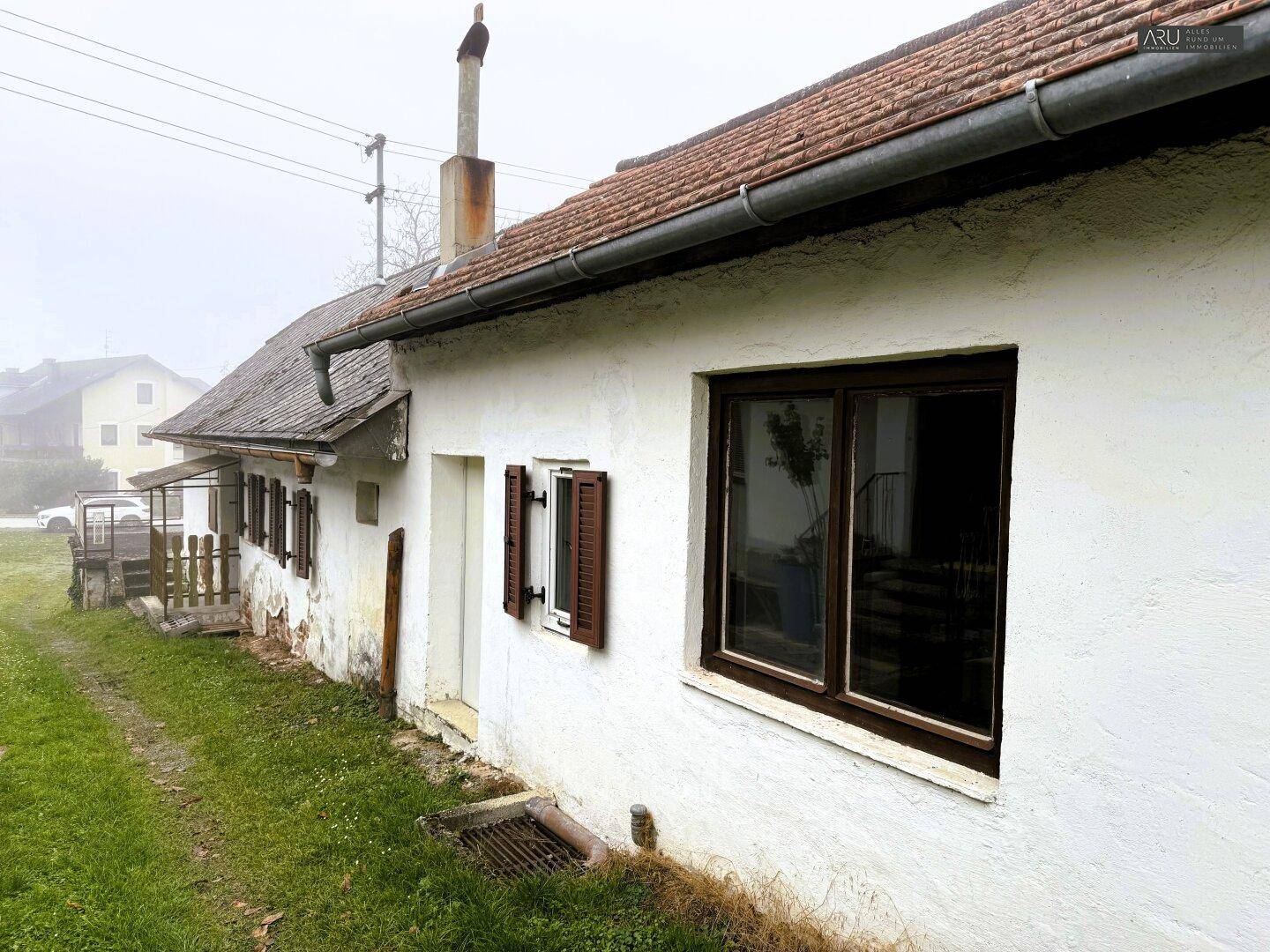 Seitenansicht eines einfachen, älteren Wohnhauses mit braunen Fensterläden und einem Schornstein.