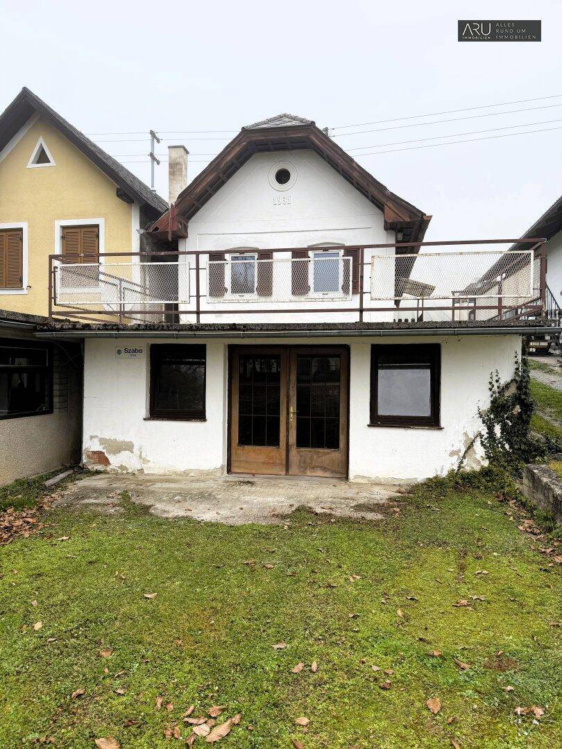 Rückseite eines älteren Wohnhauses mit Balkon und einem kleinen, ungepflegten Gartenbereich.
