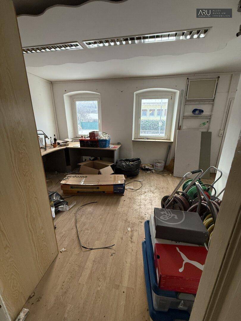 Unaufgeräumtes Büro mit zwei Fenstern, Holzboden und verschiedenen Gegenständen.