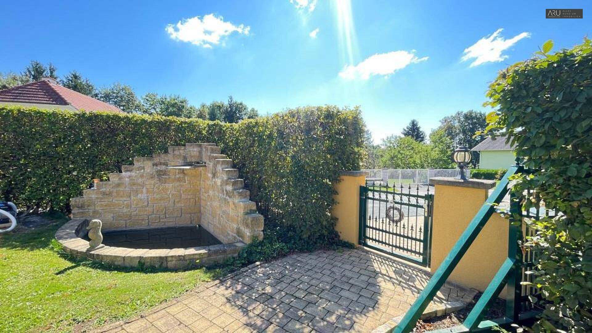 Gartenbereich mit dekorativem Steinbrunnen, grüner Hecke und schmiedeeisernem Gartentor.