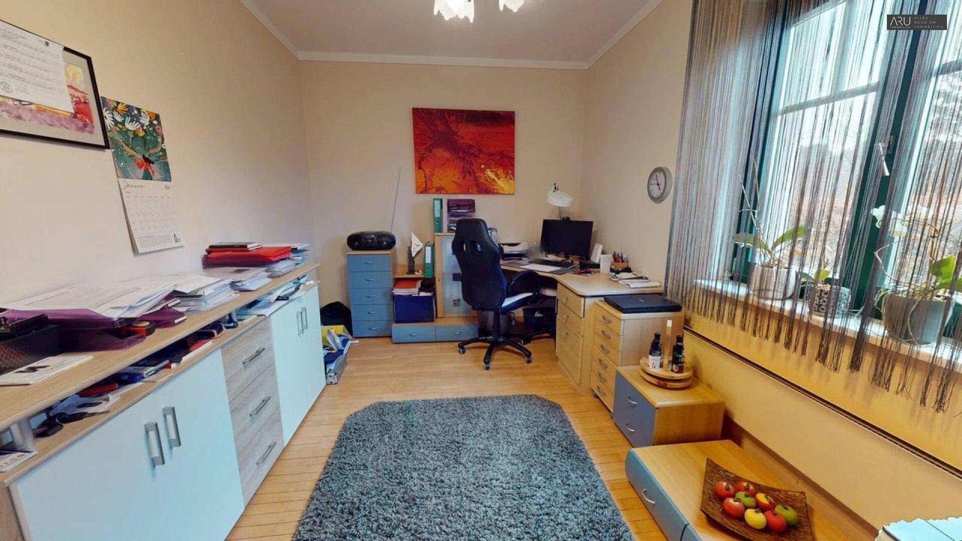 Helles Arbeitszimmer mit Schreibtisch, Regalen und Blick ins Grüne durch das Fenster.