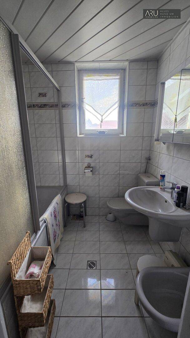 Badezimmer mit hellgrauen Fliesen, Badewanne mit Duschfunktion, WC, Waschbecken und Fenster.