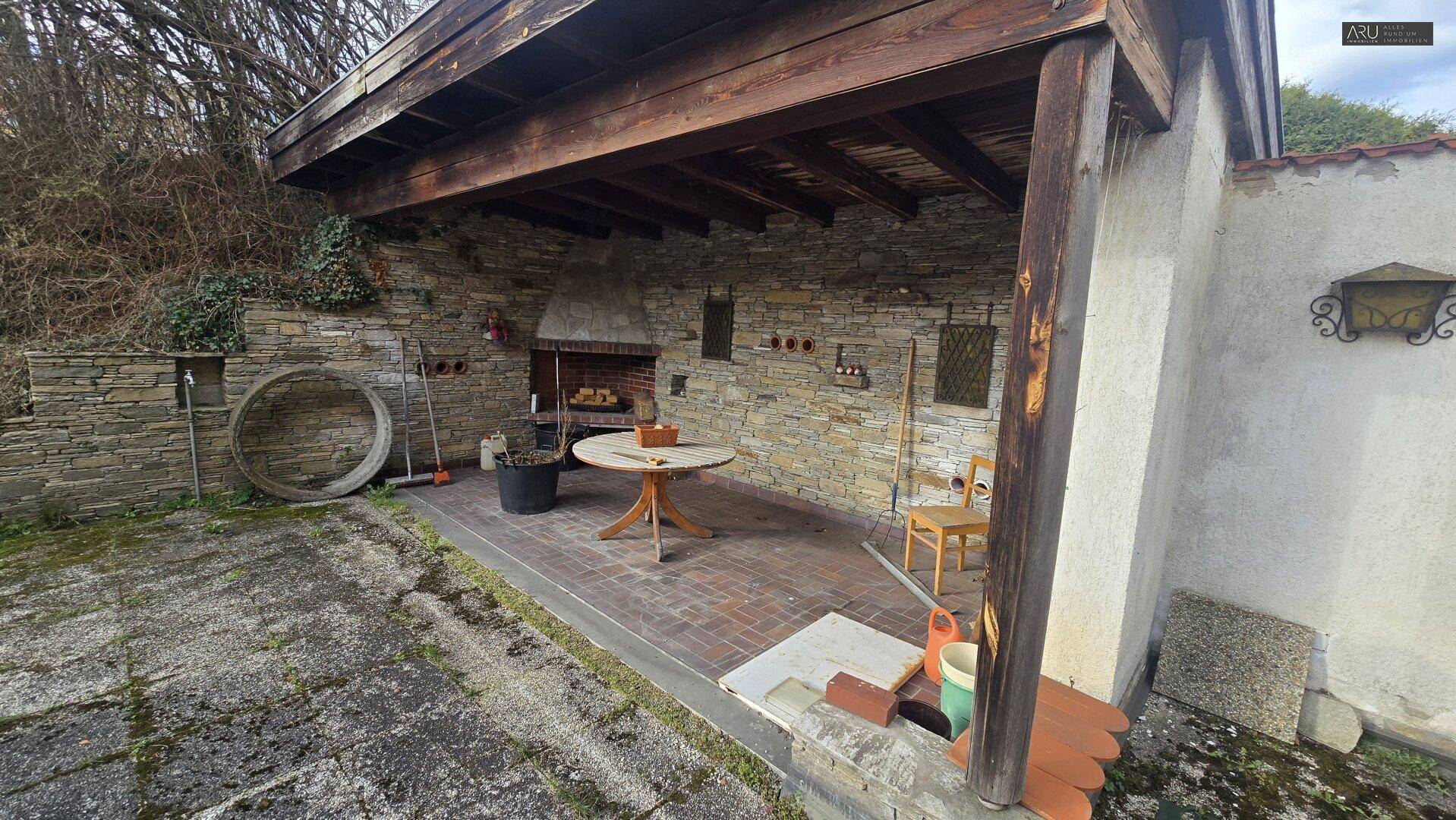Überdachter Außenbereich mit Steinmauer, integriertem Kamin oder Ofen und Holztisch.