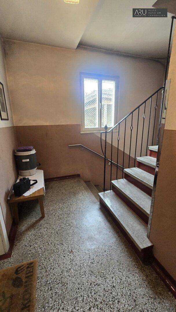 Treppenhaus mit Terrazzo-ähnlichem Boden, Metallgeländer und einem Fenster, das Licht spendet.