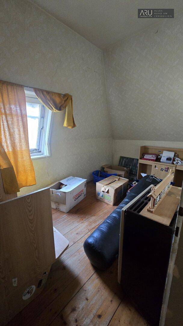 Dachbodenraum mit schrägen Wänden, Holzboden und einem Fenster, genutzt als Abstellfläche.