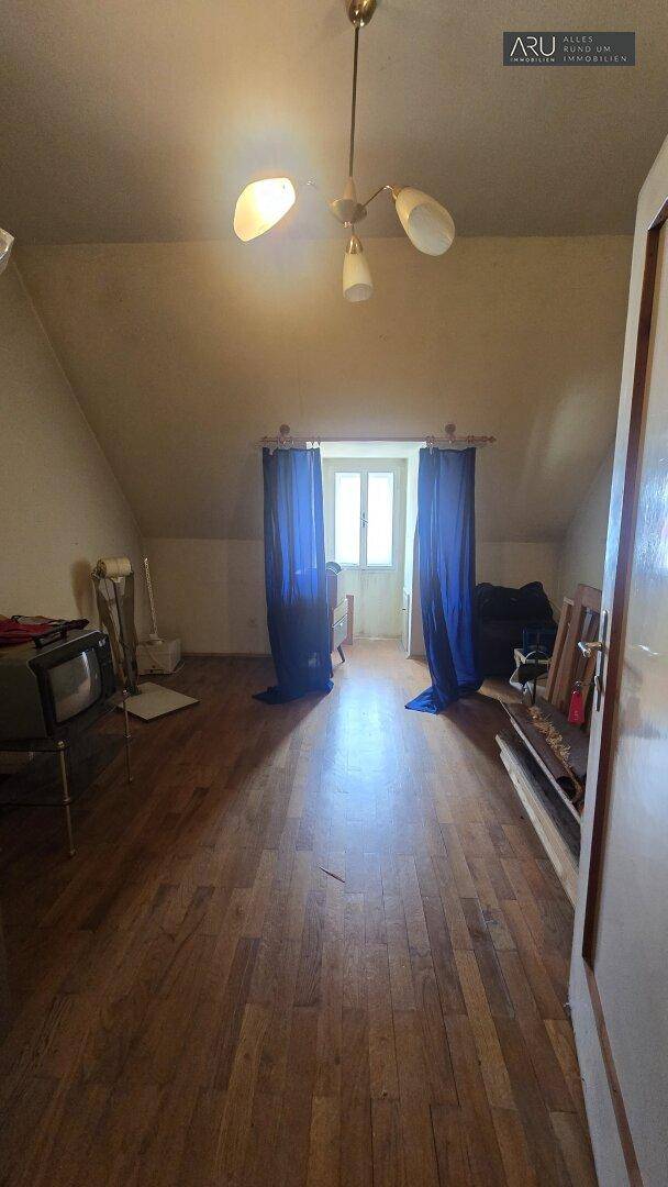 Geräumiger Wohnraum im Dachgeschoss mit Holzboden, Dachschrägen und einem Fenster mit blauen Vorhängen.