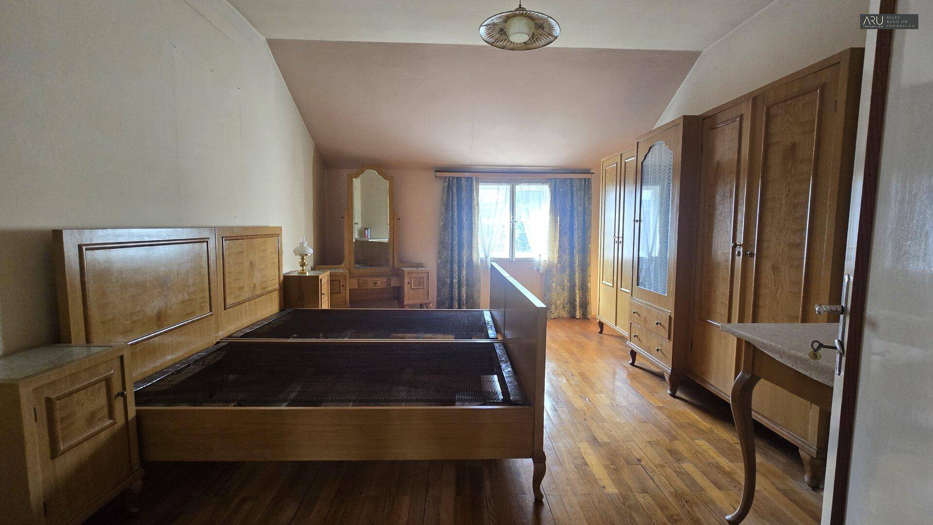 Schlafzimmer im Dachgeschoss mit großem Holzbett, Kleiderschrank und Fenster mit blauen Vorhängen.