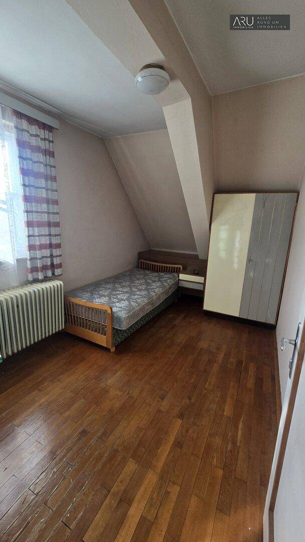 Schlafzimmer im Dachgeschoss mit Holzboden, Einzelbett, Heizkörper und Fenster mit karierten Vorhängen.