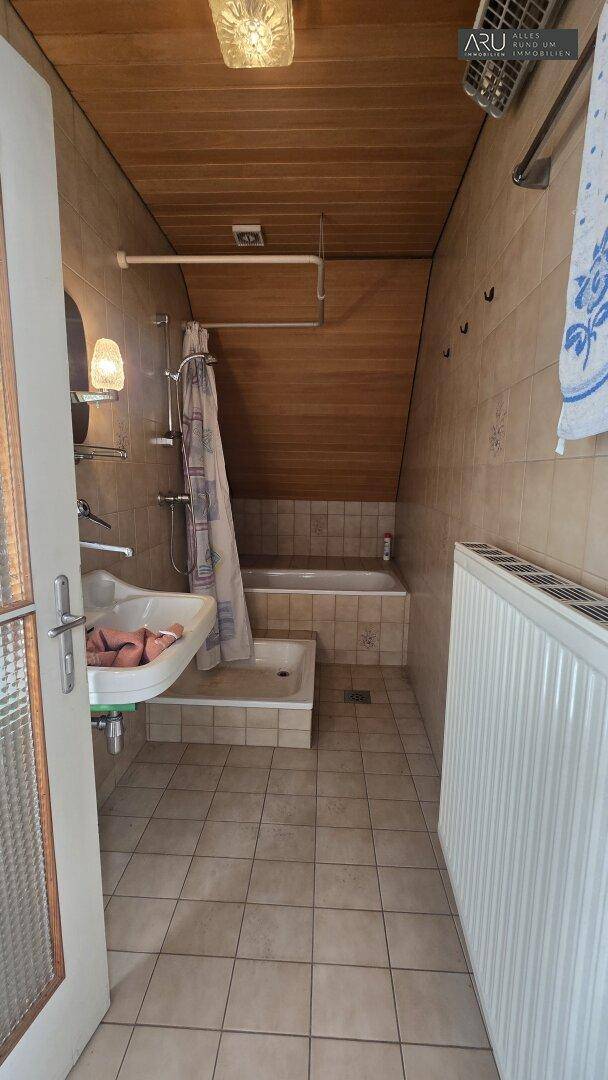 Badezimmer mit Fliesenboden, Holzdecke, Badewanne mit Duschvorhang und Heizkörper unter der Dachschräge.