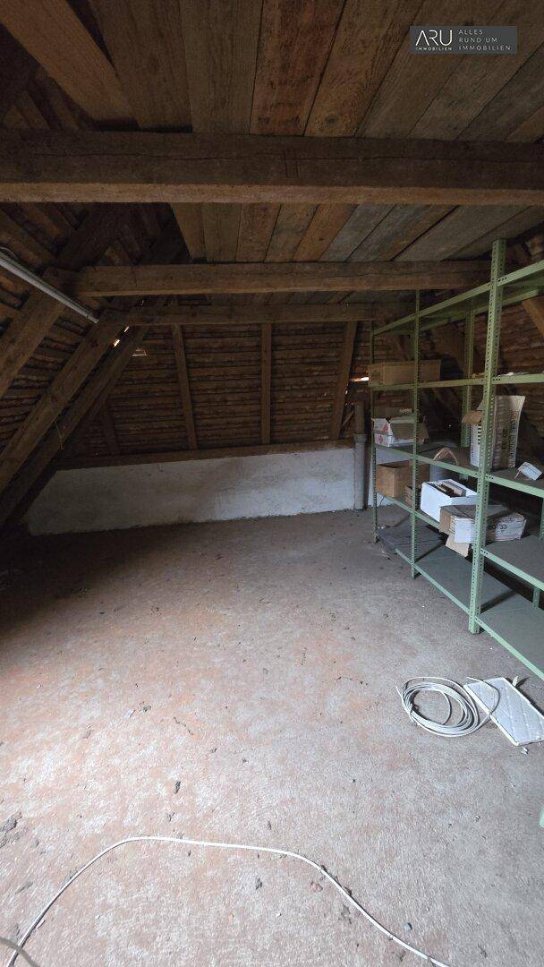 Weiterer Blick in den unausgebauten Dachboden mit Holzbalken und einem Metallregal für Lagerung.