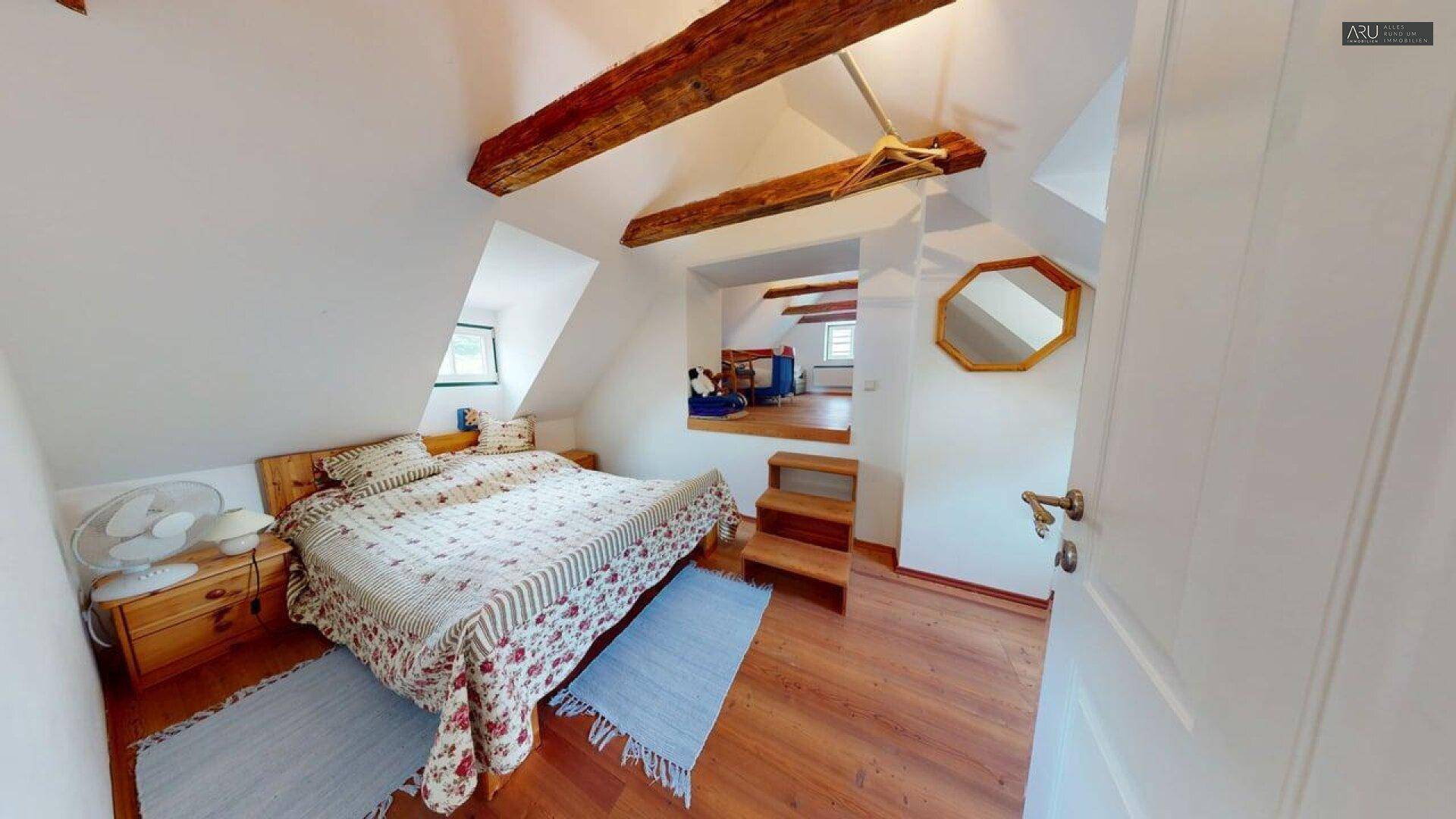 Helles Dachgeschoss-Schlafzimmer mit Holzbalken, Holzboden und Zugang zu einem weiteren Bereich.