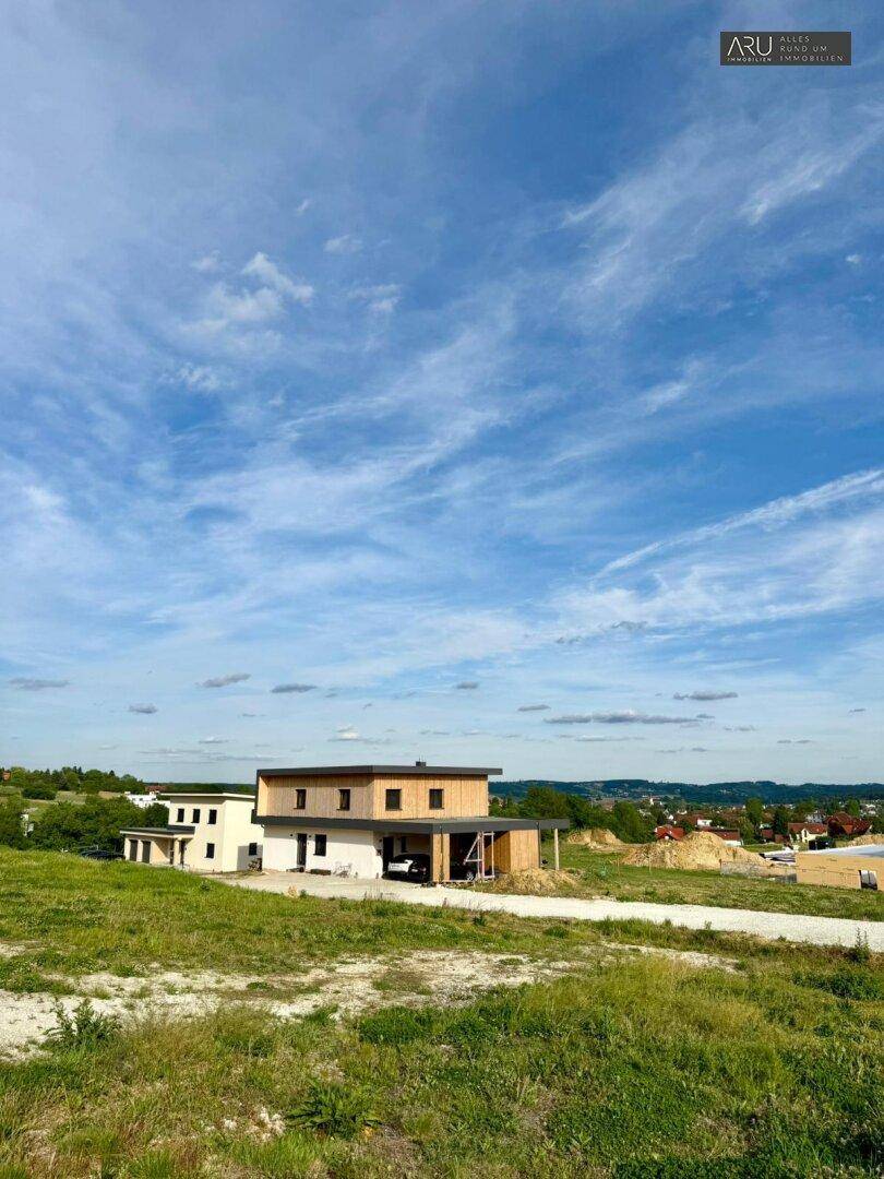 Modernes Haus mit Holzfassade und Flachdach, umgeben von grünen Feldern und Baustellen.