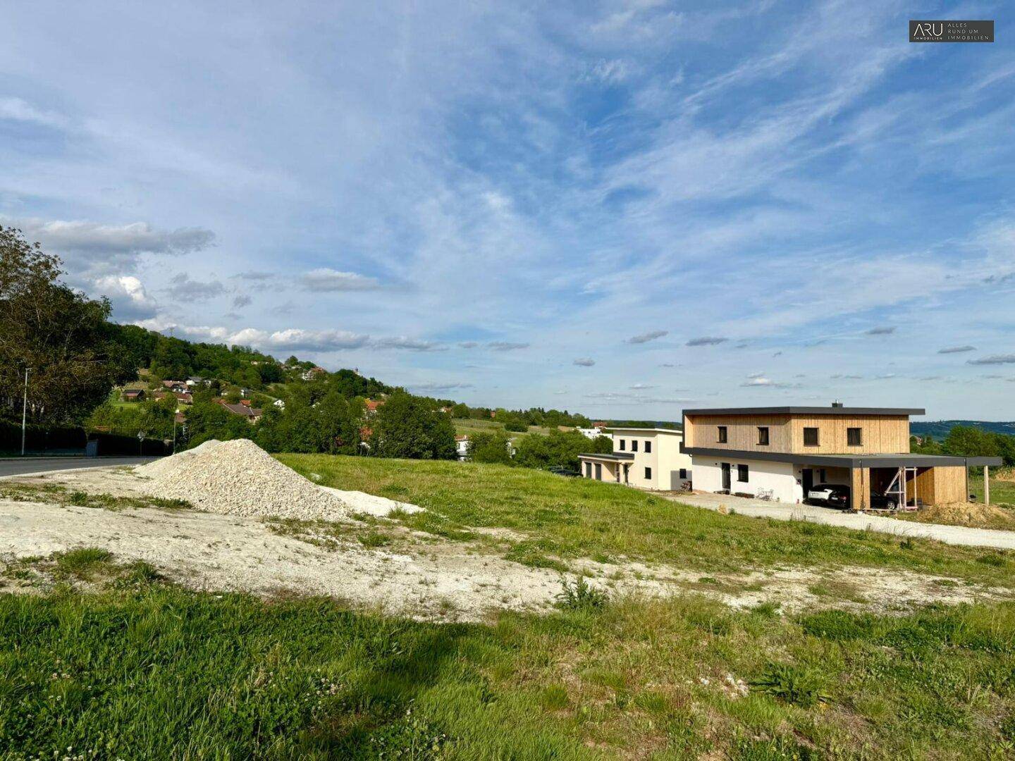 Grüne Wiese und Kieshaufen im Vordergrund, mit modernen Häusern und hügeliger Landschaft im Hintergrund.