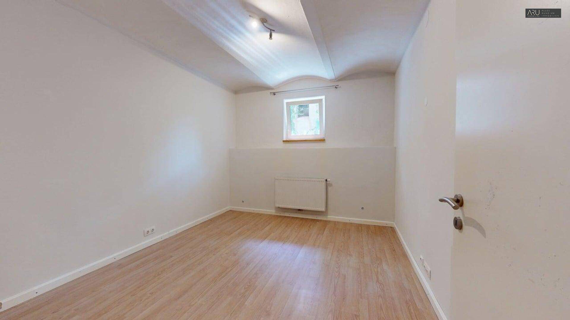 Schlichtes Zimmer mit Laminatboden und einem Fenster, das Tageslicht hereinlässt.