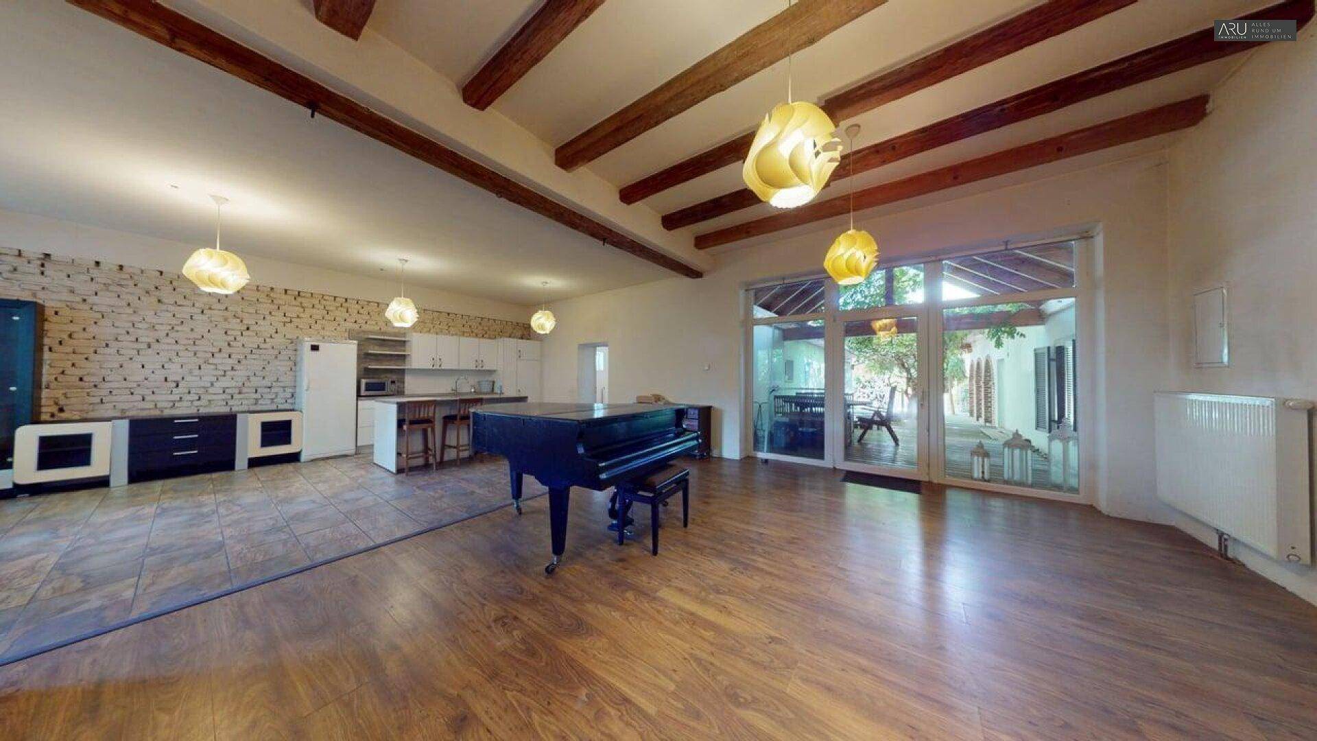 Offener Wohnbereich mit Holzbalken, Klavier und Zugang zu einem Wintergarten oder Terrasse.