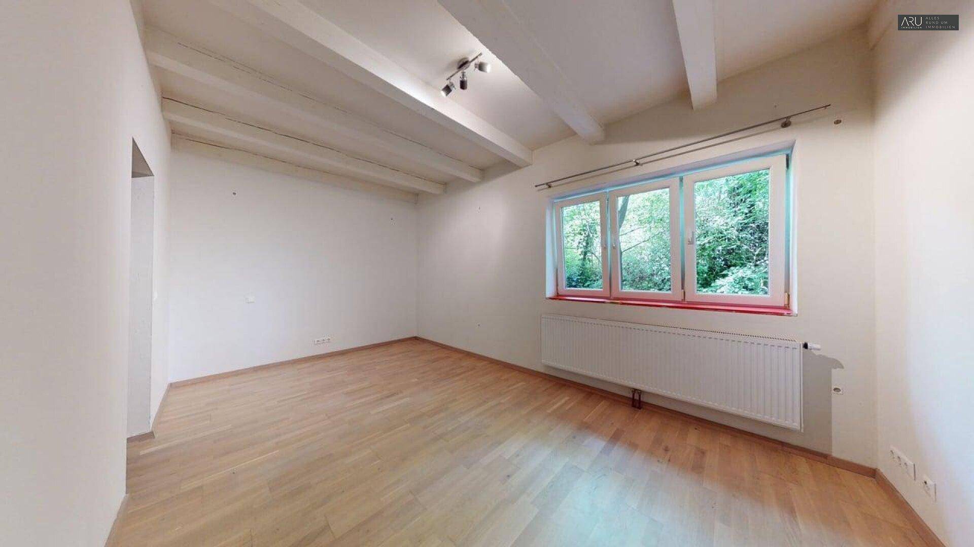 Helles, leeres Zimmer mit Holzbalkendecke und Blick ins Grüne.