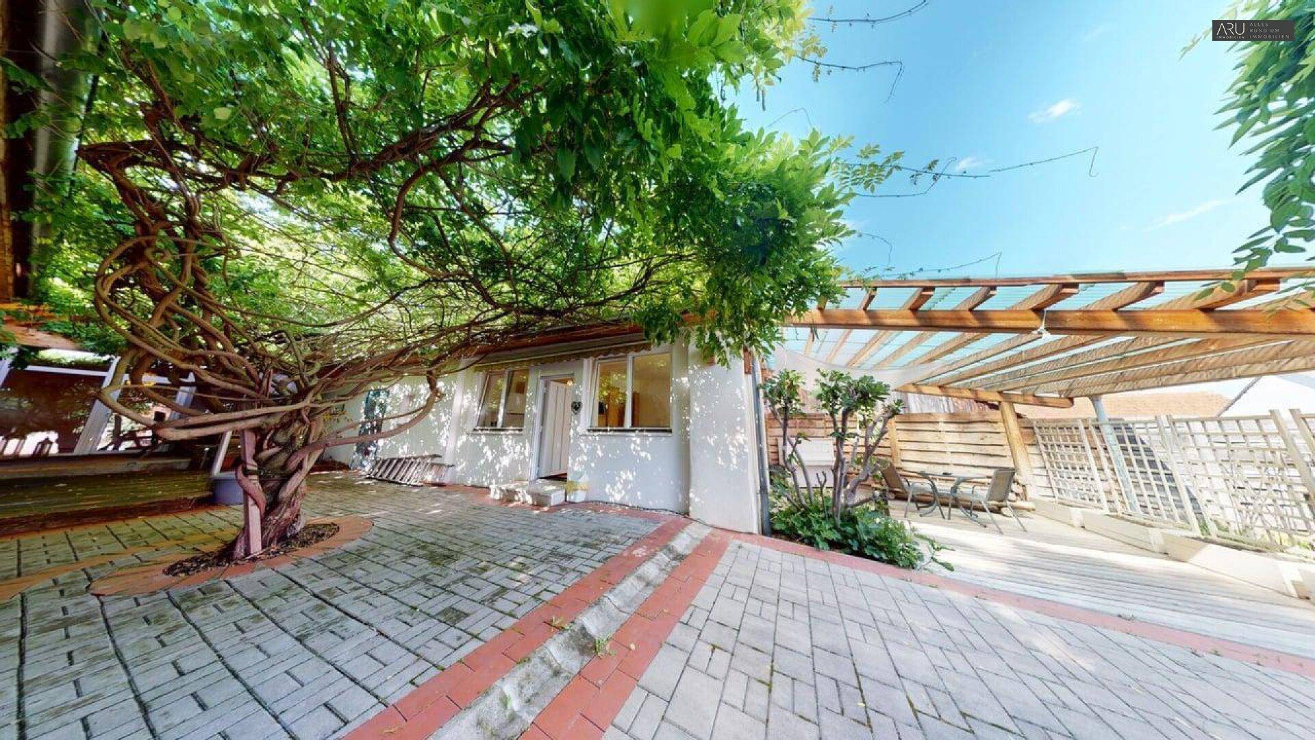 Gepflasterter Patio mit üppiger Begrünung, Pergola und Sitzgelegenheiten im Freien.