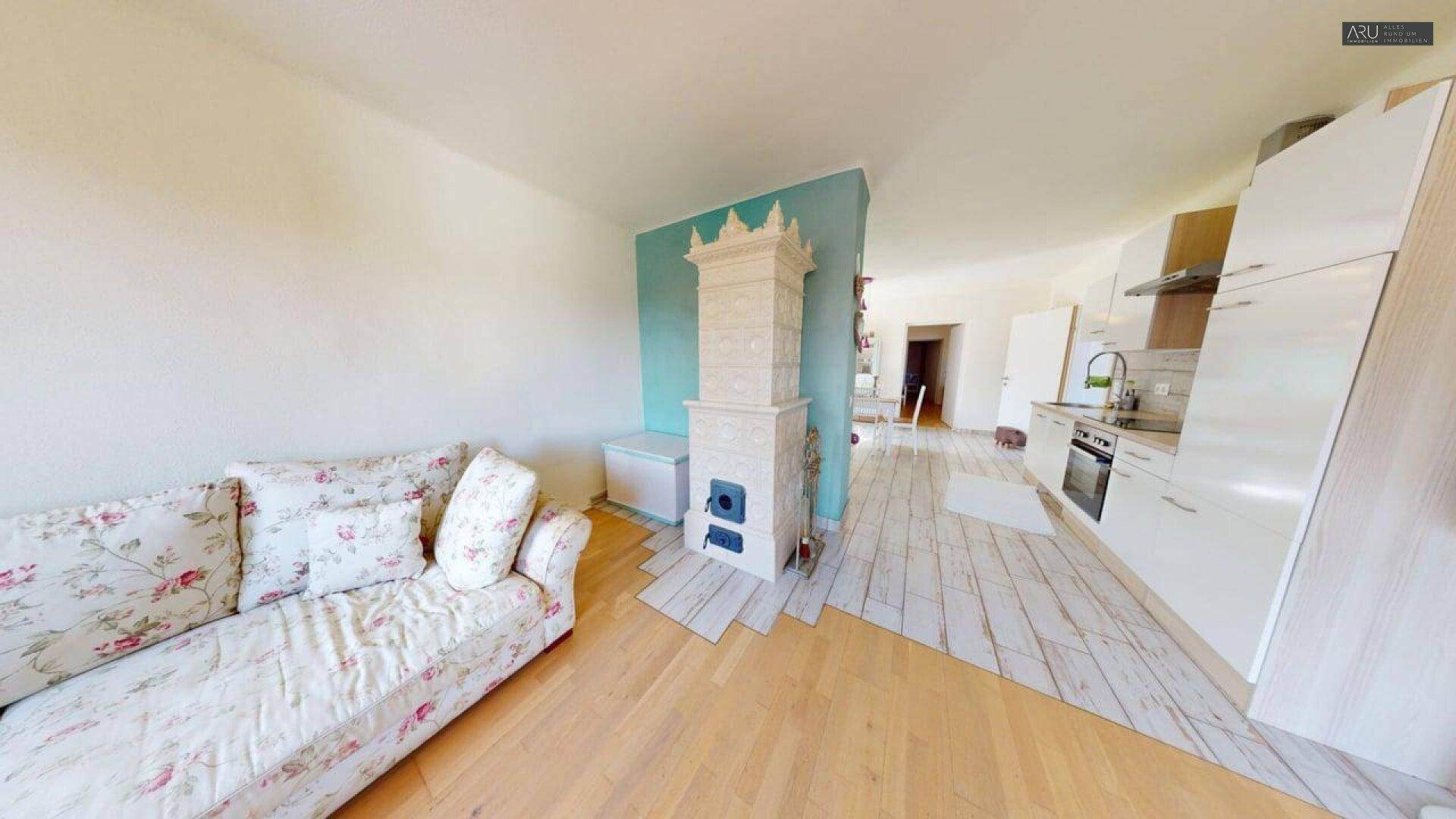 Offener Wohnbereich mit Kachelofen, Sofa und moderner Küchenzeile im Hintergrund.