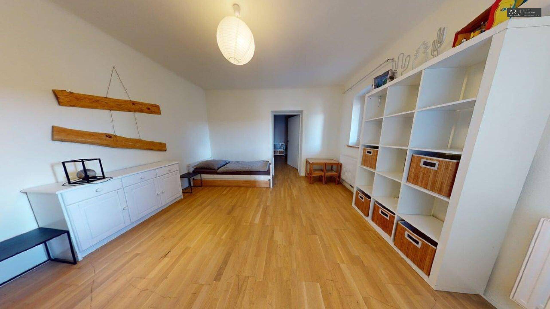 Geräumiges Zimmer mit Holzboden, weißem Regal und einfacher Einrichtung.