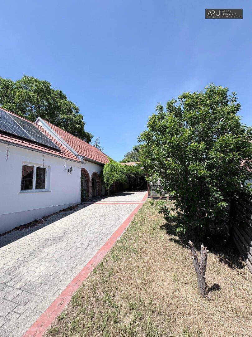Gepflasterte Zufahrt neben dem Haus mit Solarpaneelen auf dem Dach und Bäumen.