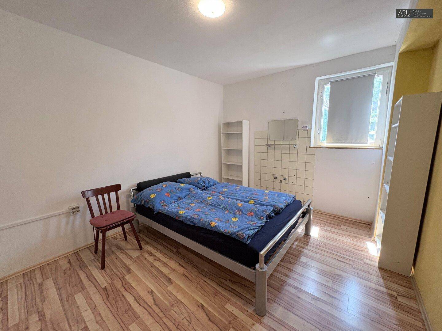 Einfaches Schlafzimmer mit Doppelbett, Regalen und einem kleinen Waschbereich.