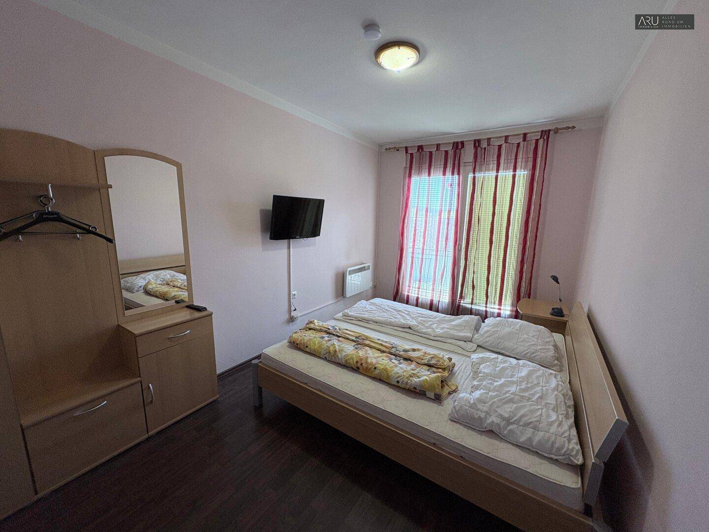 Helles Schlafzimmer mit Doppelbett, Fernseher und einfacher, funktionaler Einrichtung.