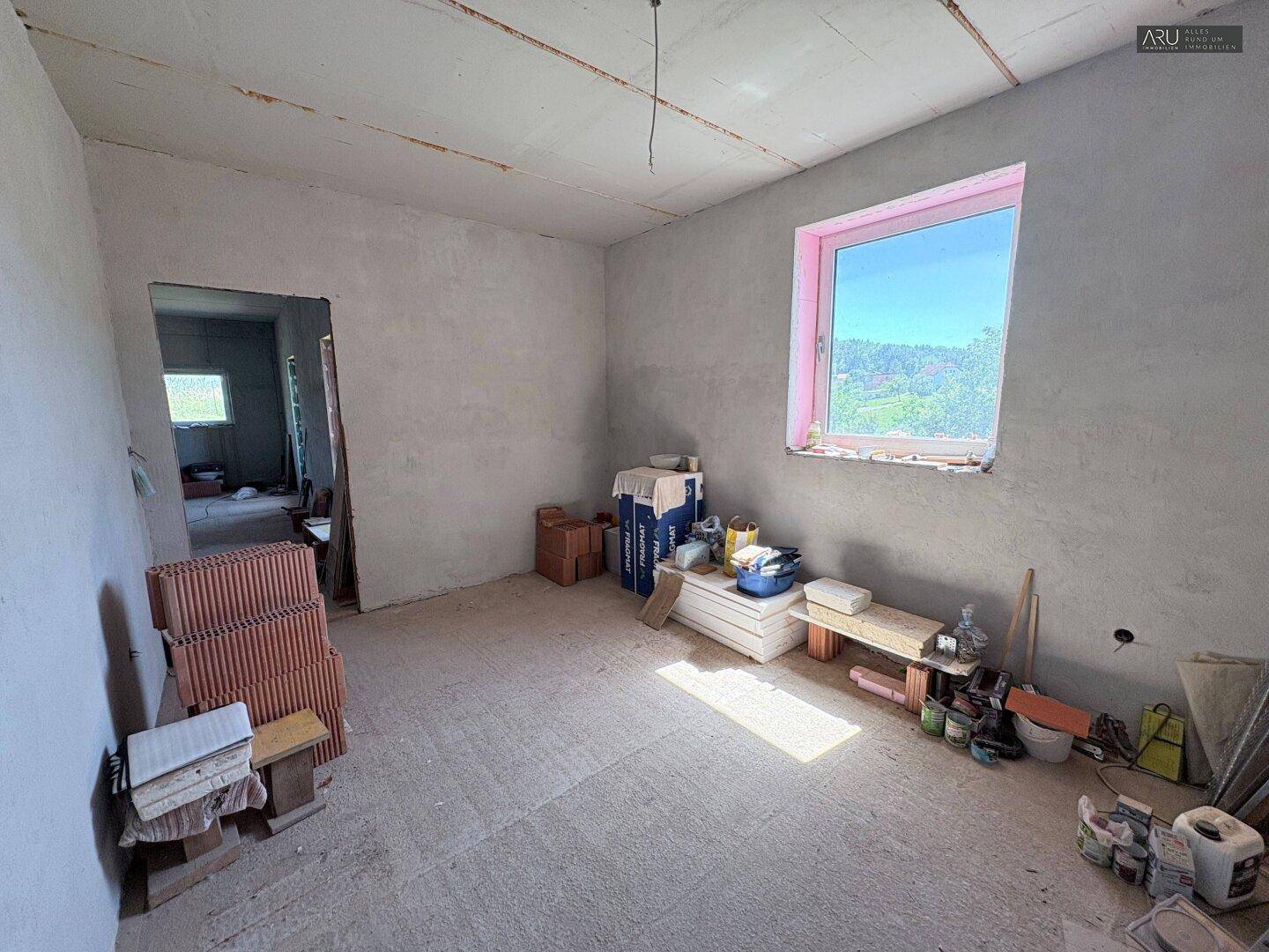 Unfertiger Raum mit grauen Wänden, Betonboden und einem Fenster mit Blick auf die Landschaft.