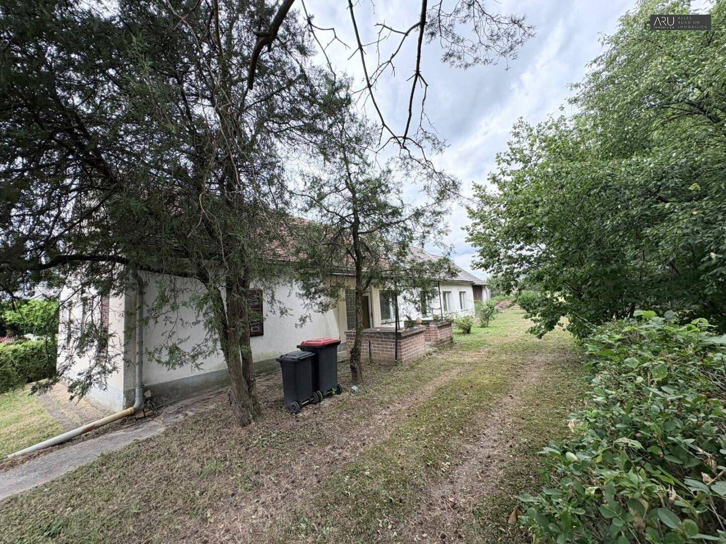 Ländliches Haus mit Zufahrt, Mülltonnen und umgebender Vegetation.