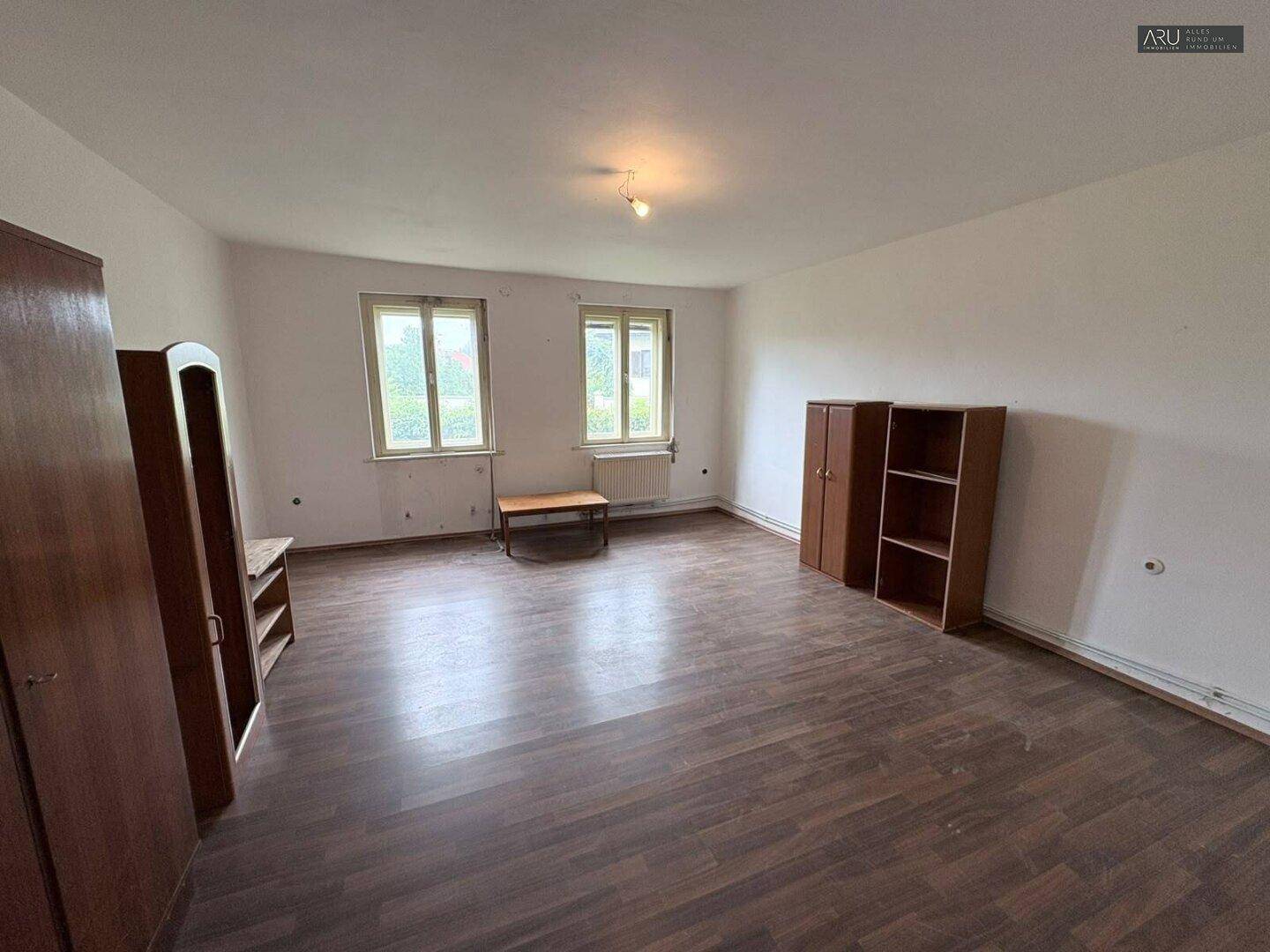 Geräumiger, unmöblierter Raum mit dunklem Laminatboden und zwei Fenstern.