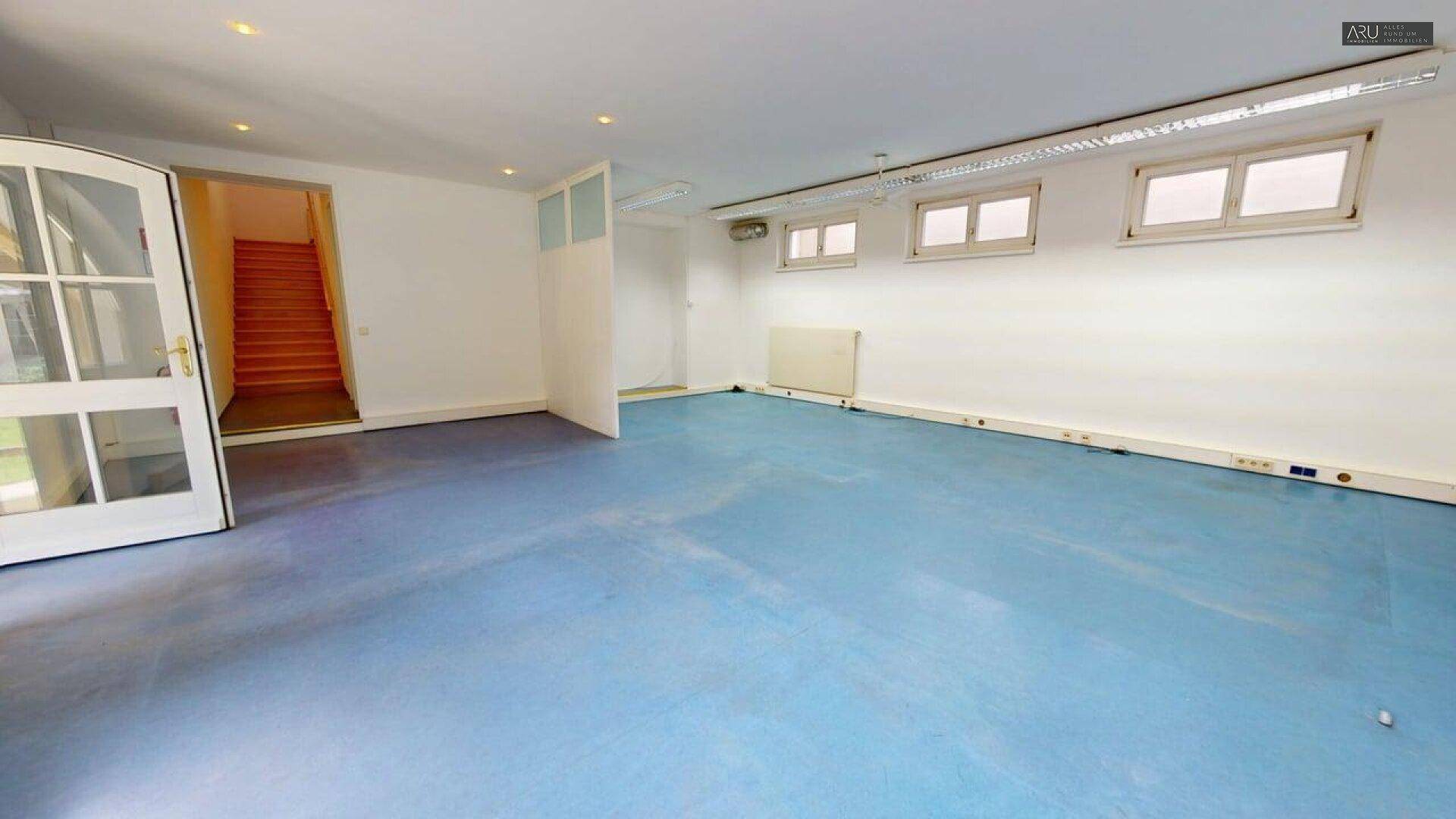 Großer, leerer Raum mit blauem Boden, weißen Wänden und hohen Fenstern, vielseitig nutzbar.