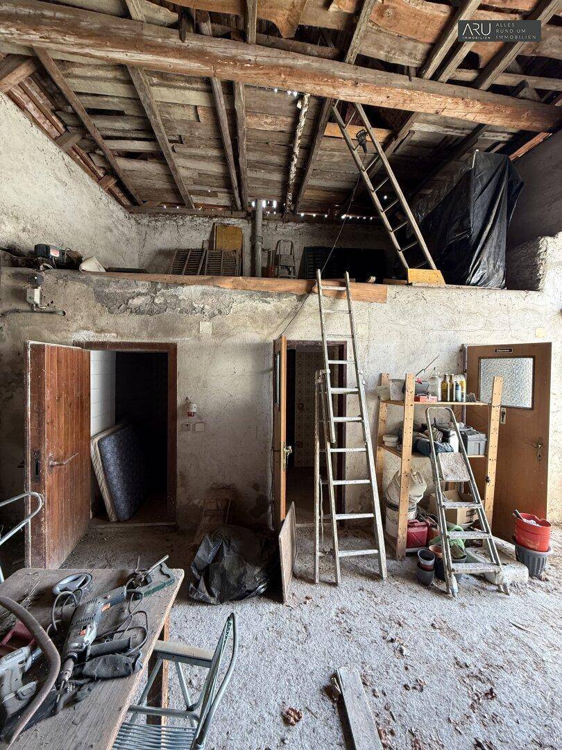 Rustikaler Innenraum mit Holzbalken, Leitern und Werkzeugen, der auf eine obere Ebene führt.