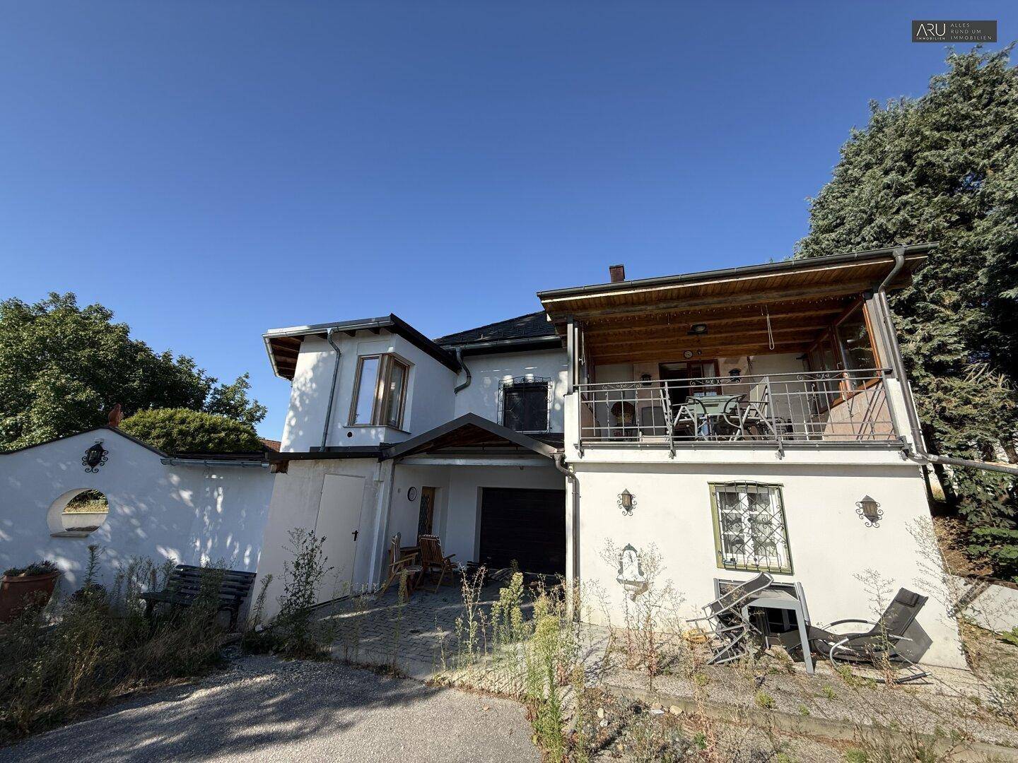 Hausfassade mit überdachtem Eingang, Garage und möbliertem Holzbalkon in sonniger Lage.